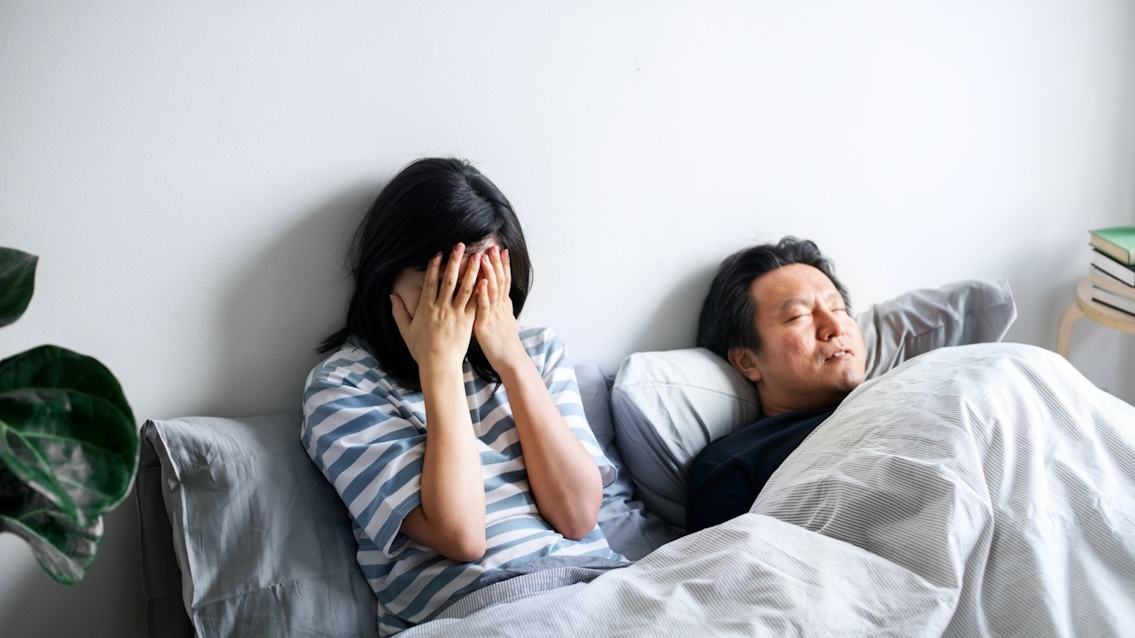 A stressed woman with hands on face siitin in bed with a man sleeping beside her.