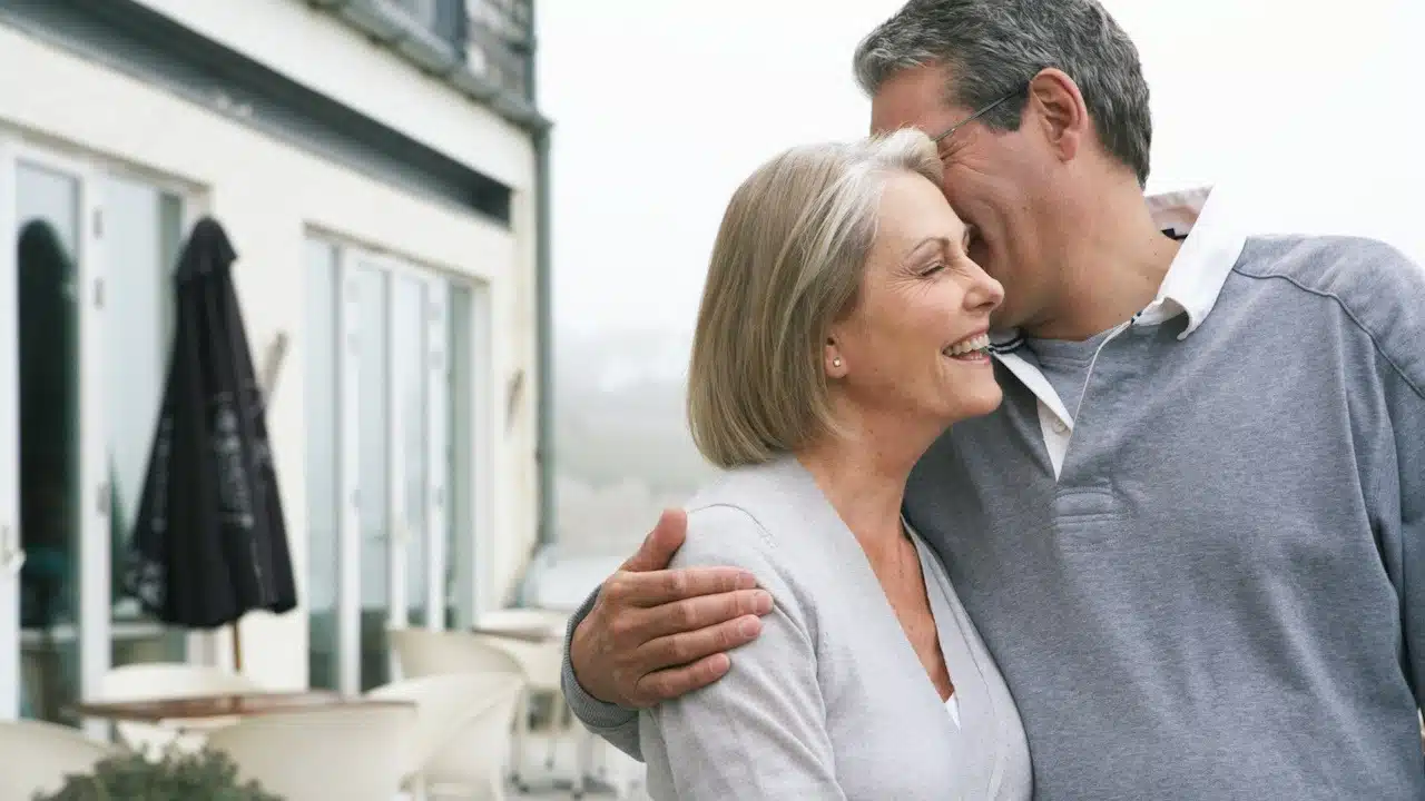 A smiling mature couple where the man is whispering something in his wife’s ear.