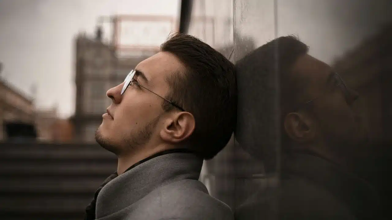 A serious man with glasses and a beard is leaning his head against a stone wall, looking up.