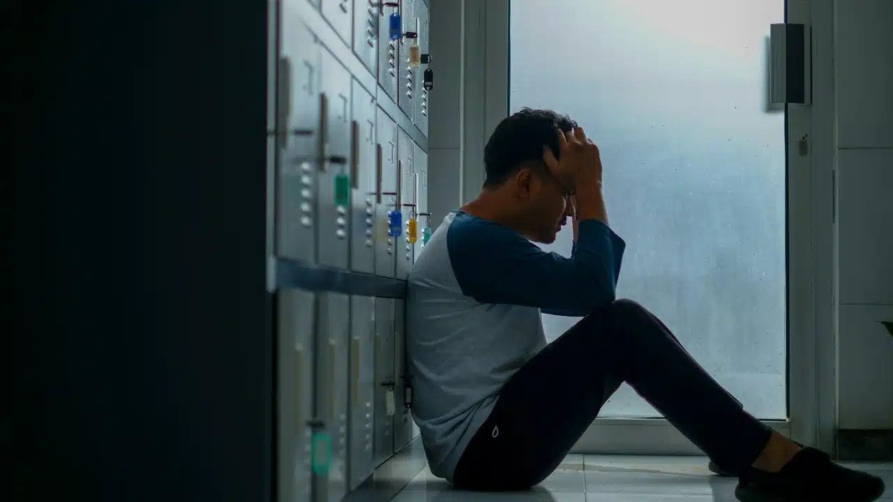 A man sitting on the floor while leaning on some lockers while holding his head in his hands.