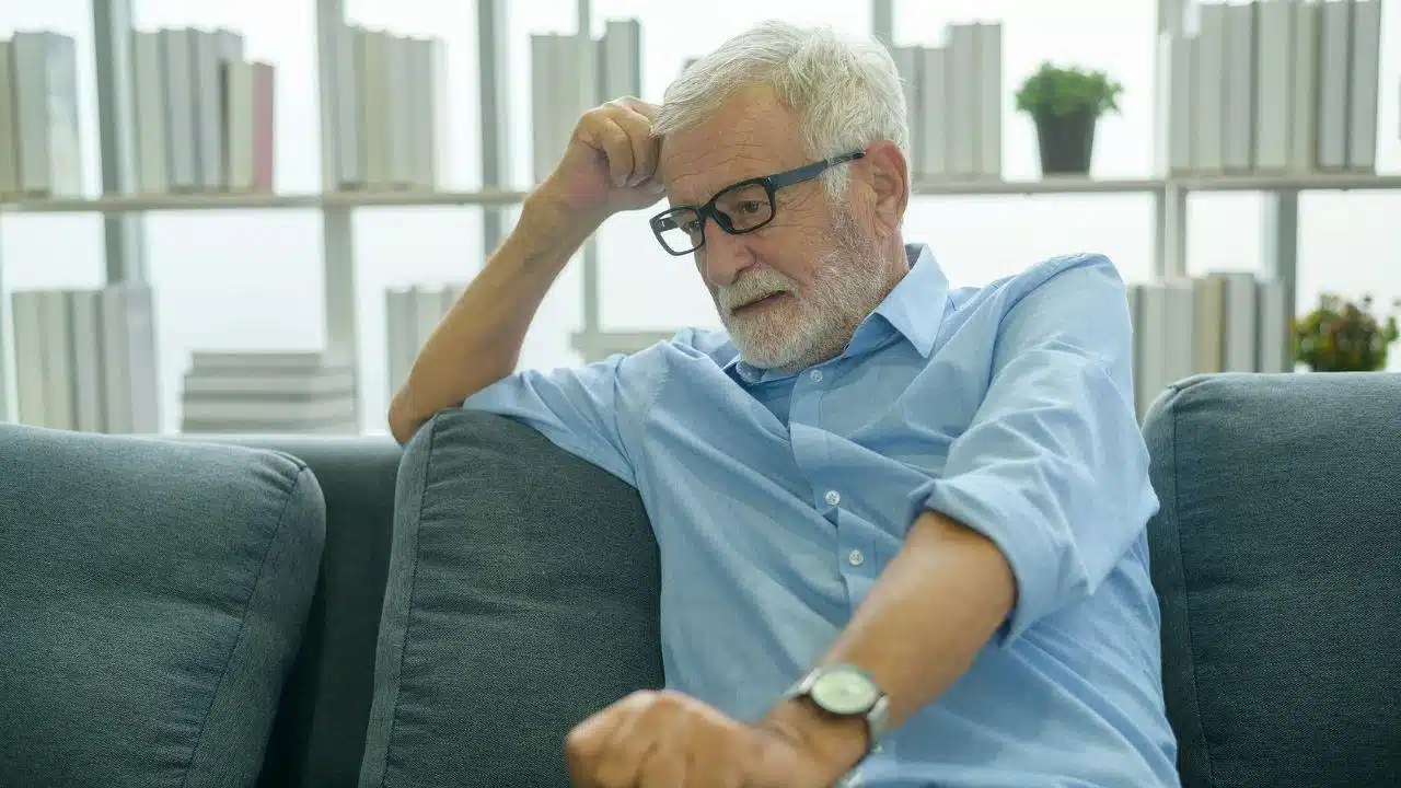 A distressed older man with glasses sits on a couch, his hand on his head.