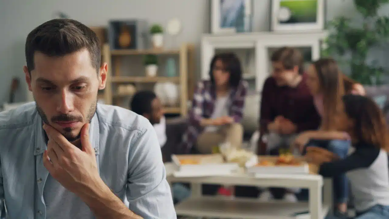 A man in the foreground looks down thoughtfully while a group of people talk behind him.