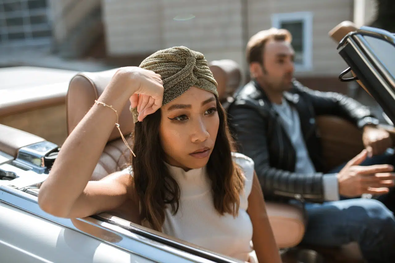 A woman in a white tank top is sitting in a car feeling annoyed while a man is driving the car.