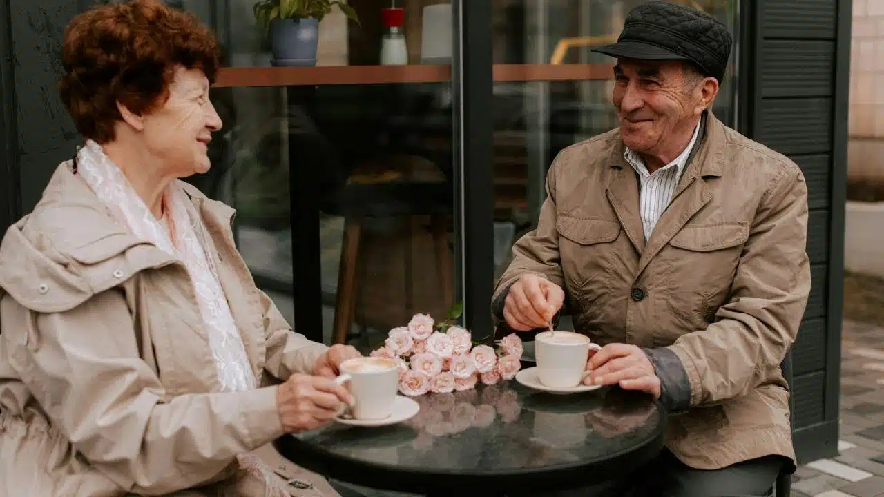 An elderly couple smiles at each other while having coffee at an outdoor table.