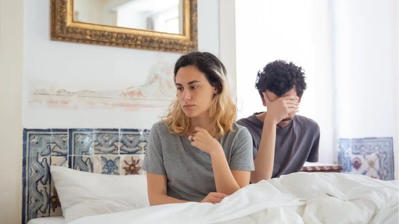 A couple sits  on a bed after an argument; the woman looks sad while the man holds his hand over his face.