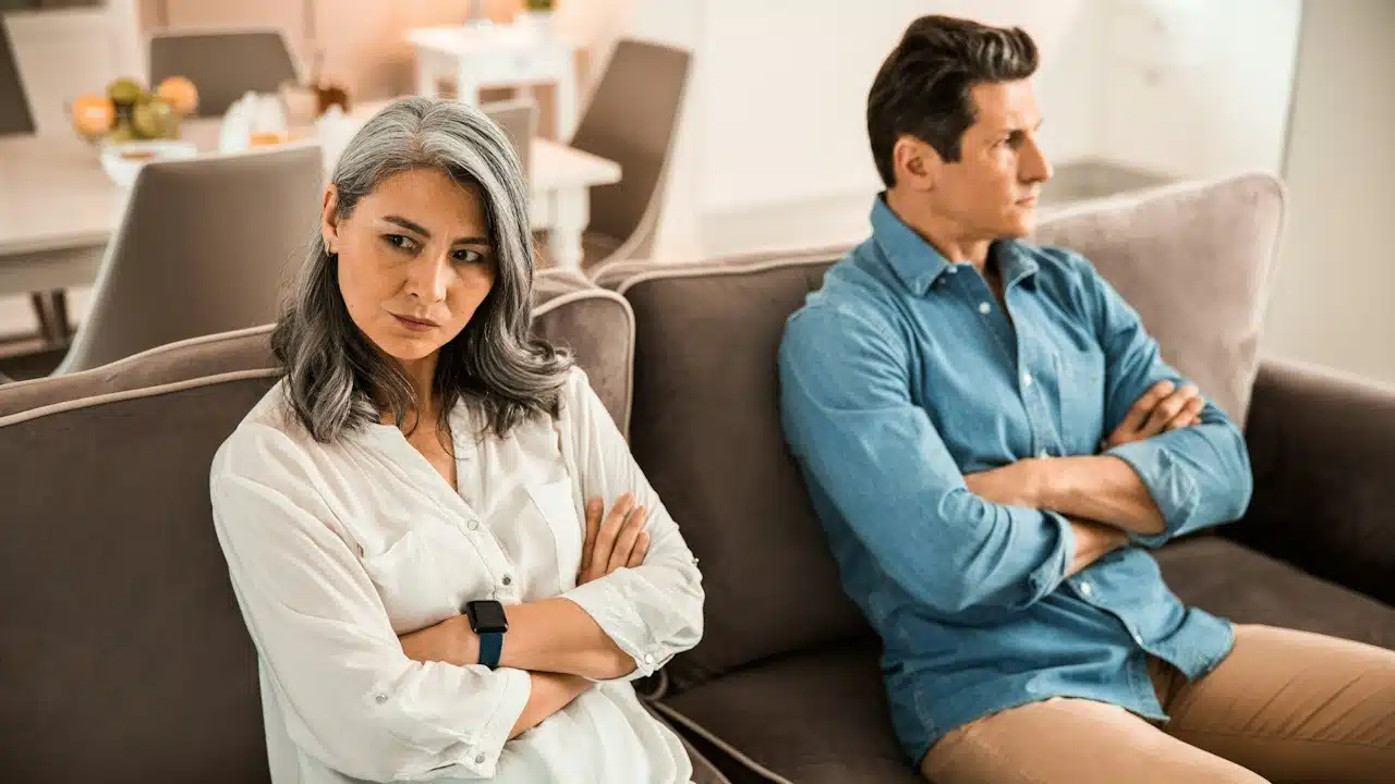 A mature older couple sitting on a couch with arms crossed and refusing to look at each other.