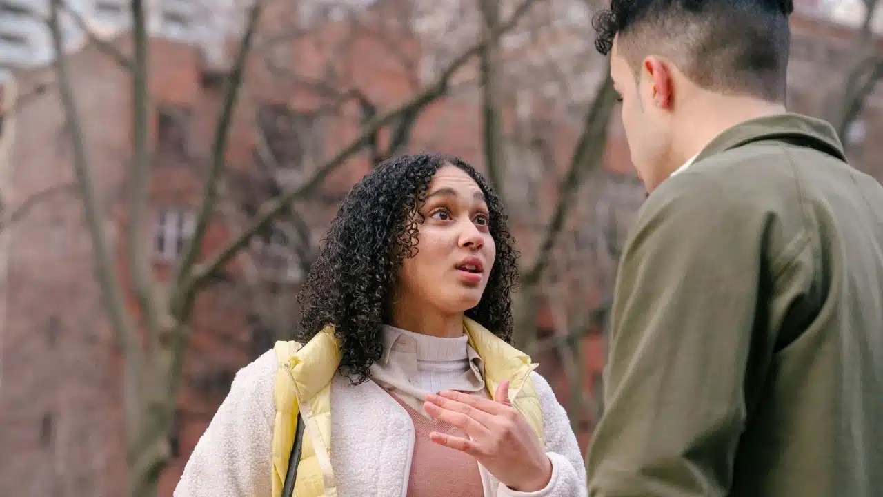 A woman with curly hair looks at a man and gestures while talking outdoors.