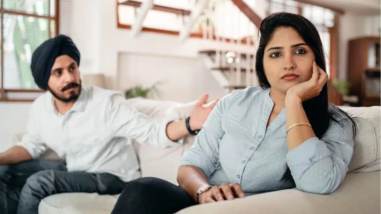 A woman is sitting on a couch with a distant expression while a man in a turban sits behind her, gesturing toward her.