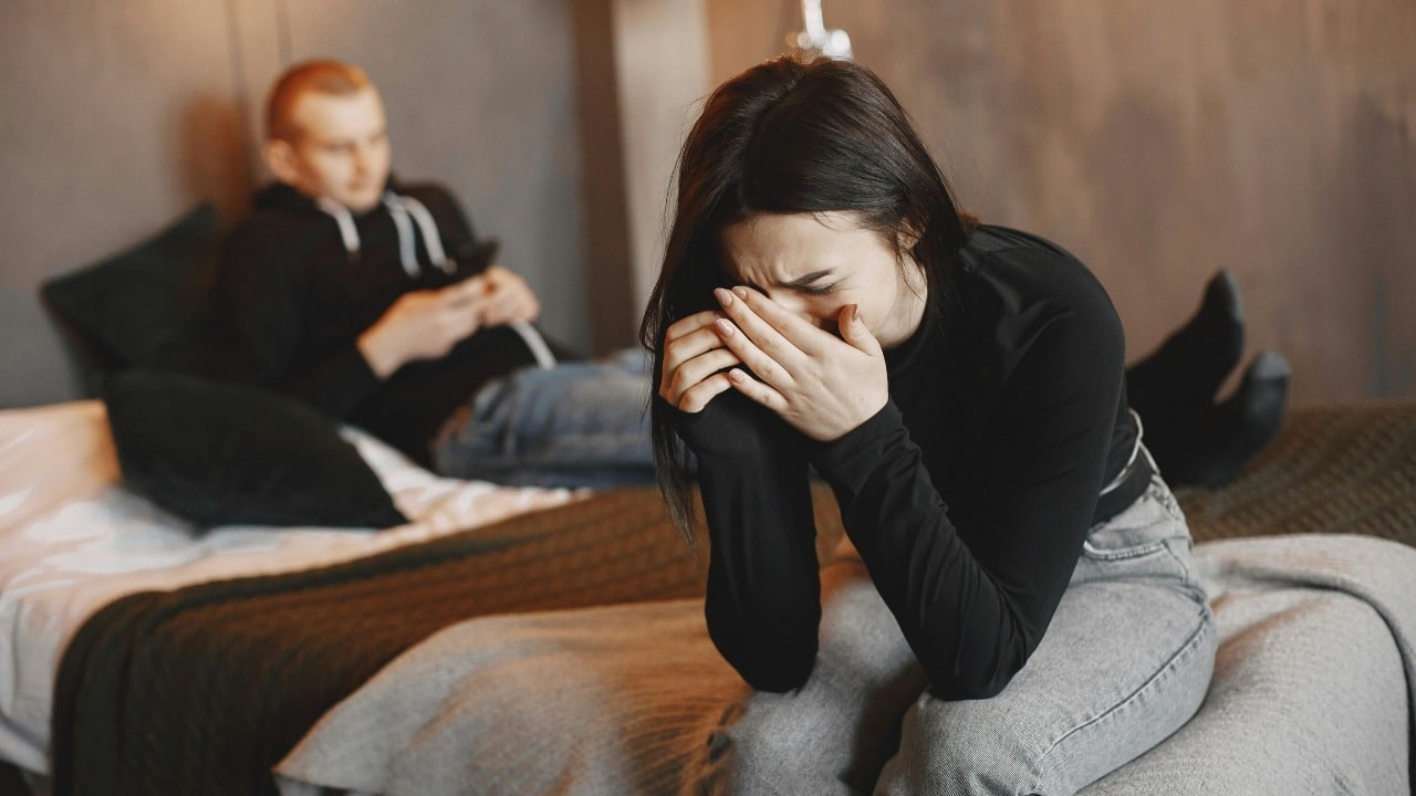 A woman sits on a bed covering her face while a man sits behind her.