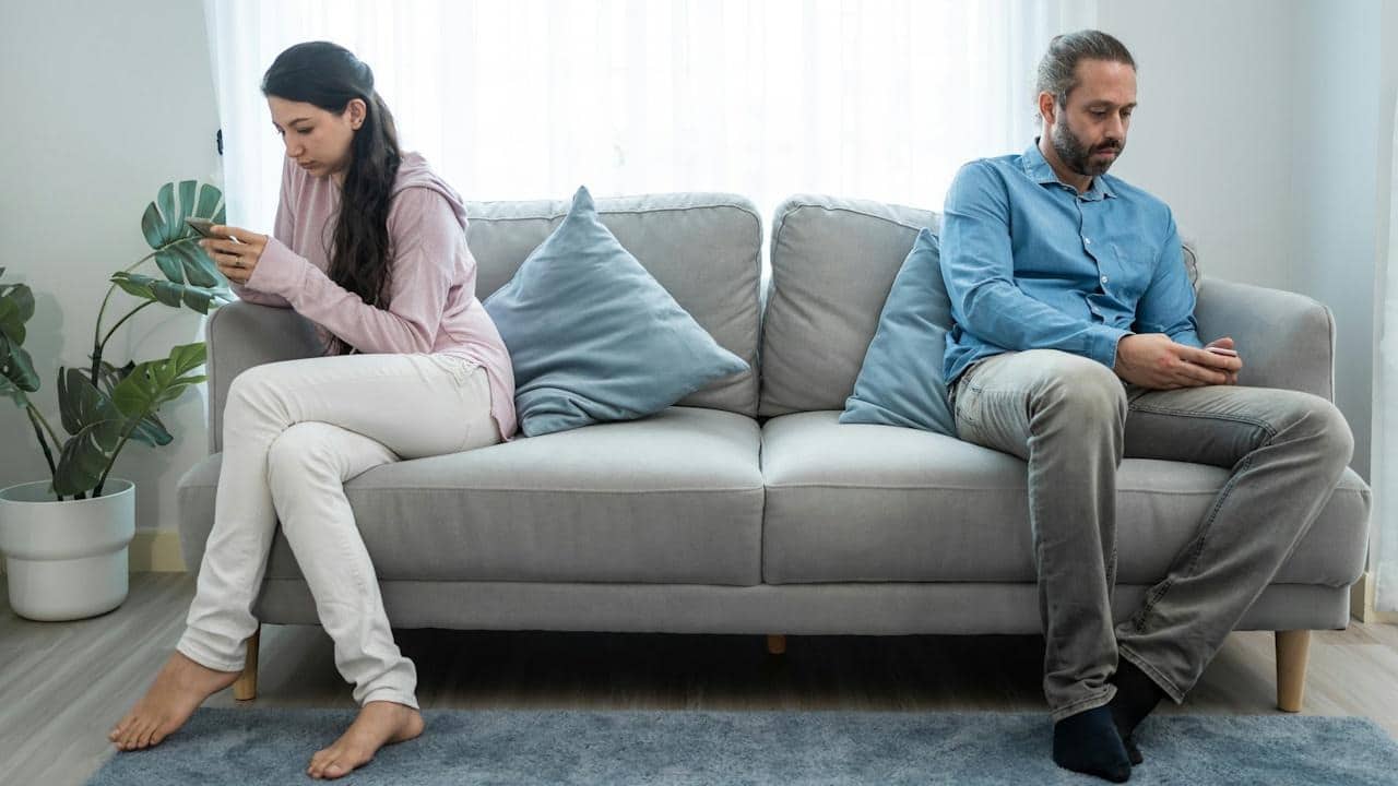 A man and a woman are sitting far apart on a couch, each looking at their phone and ignoring the other.