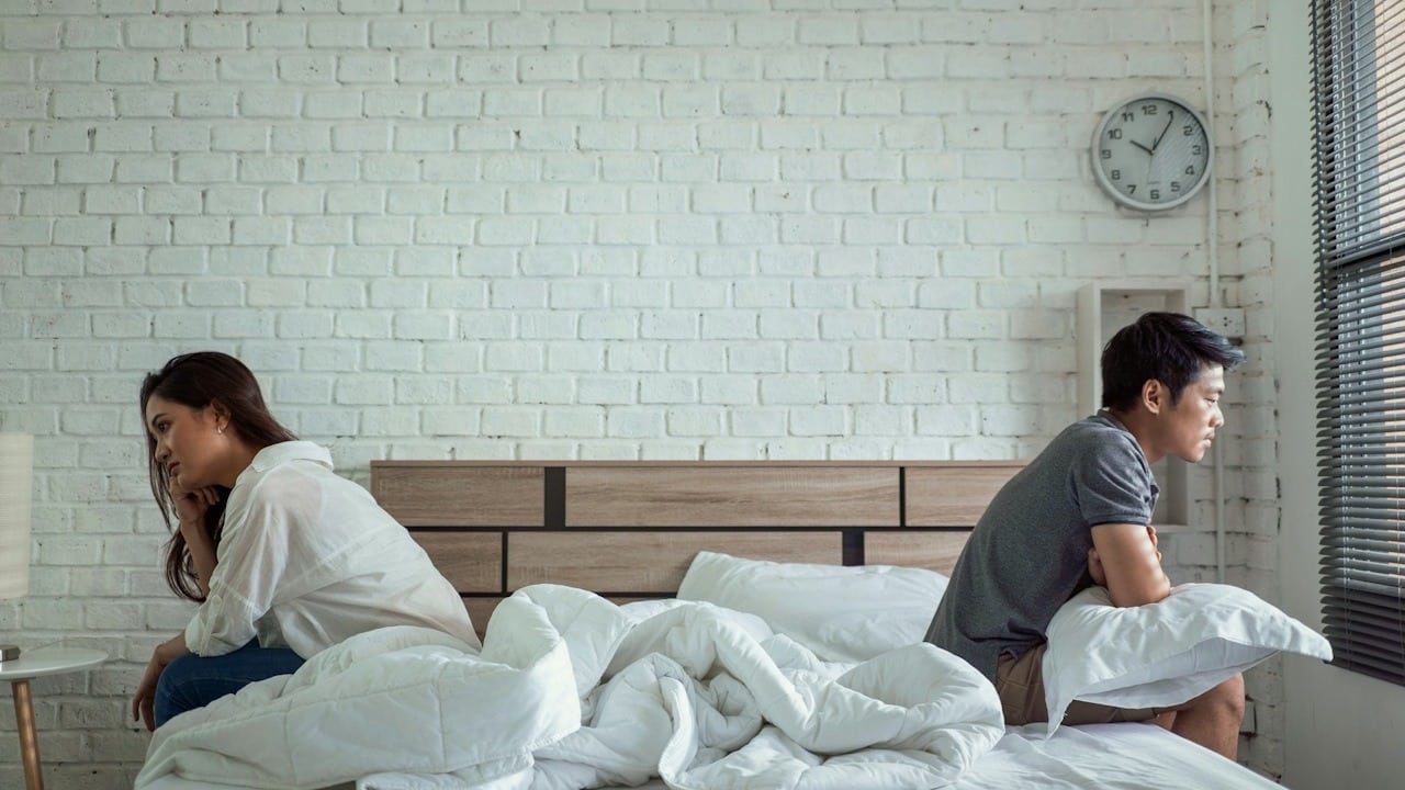 A couple sitting on separate ends of a bed facing away from each other.