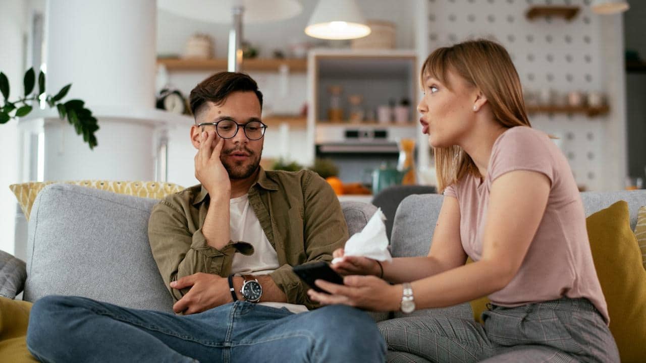 A woman is speaking emphatically to a man who looks distressed as they sit on a couch in a living room.
