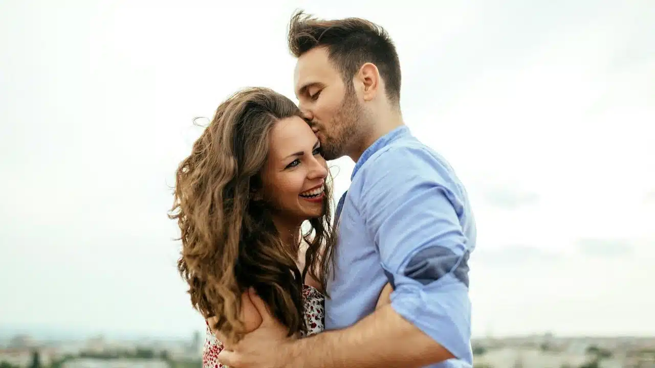 A lightly bearded man kissing a smiling woman on her temple.
