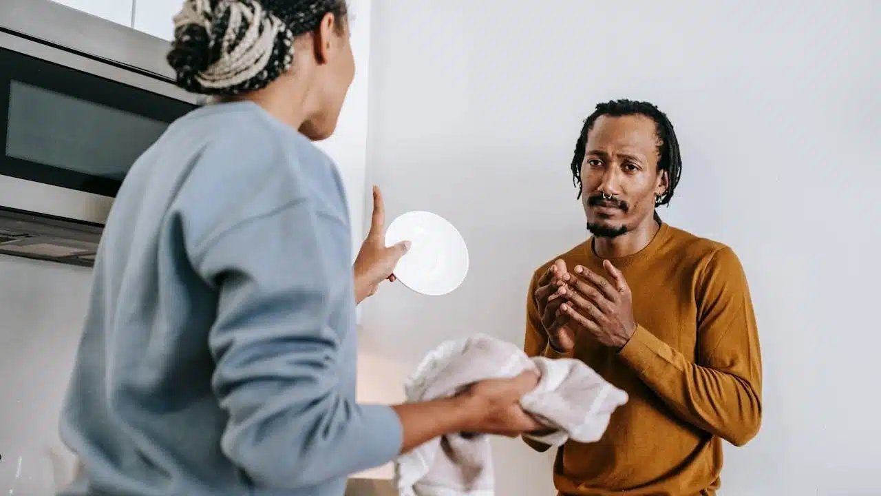A woman is holding a plate and towel, gesturing while speaking to a man who looks distressed.