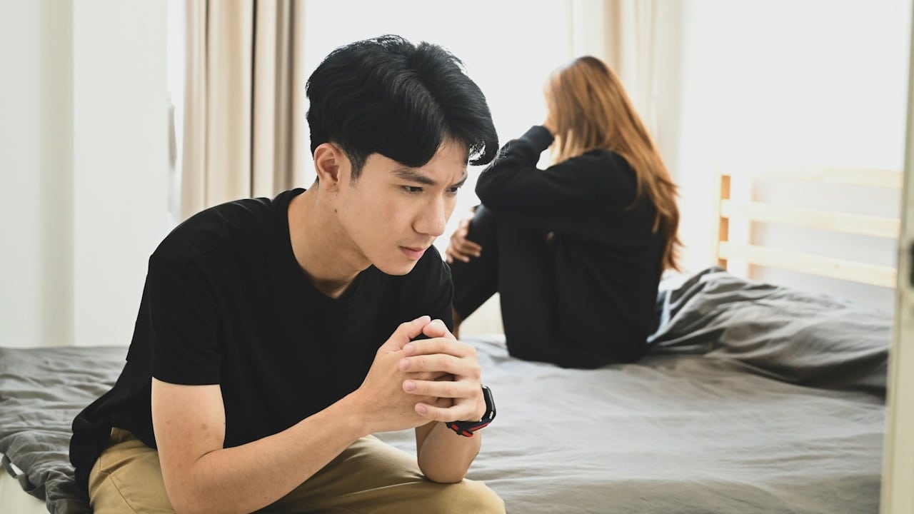 A man is thinking deeply while sitting on the edge of a bed and his partner sits on the bed in the background.