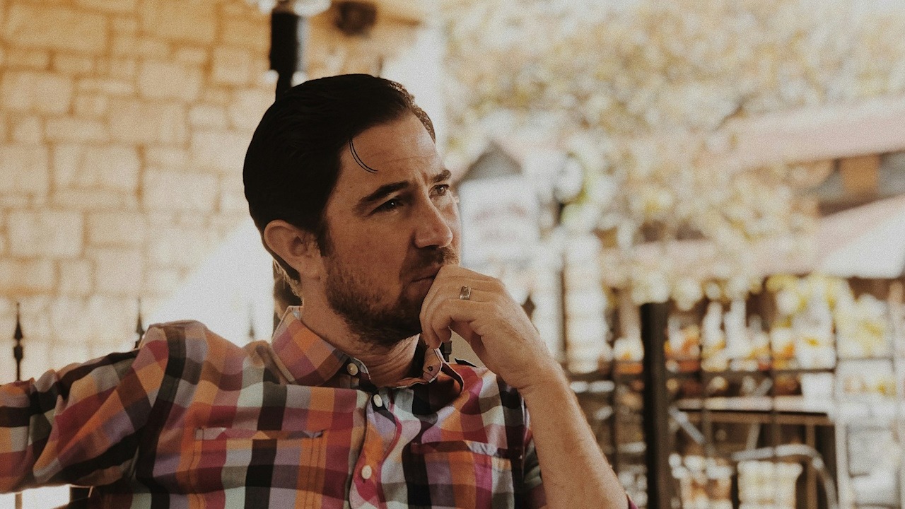 A man wearing a colored checkered shirt sitting and thinking deeply about something.