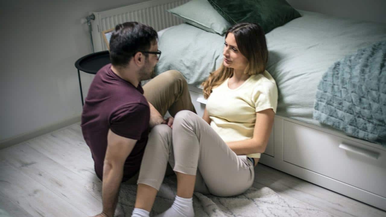 A couple conversing while sitting on the floor of their bedroom.
