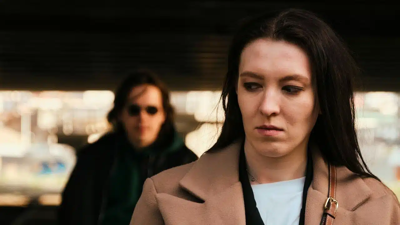 Woman in a beige coat looking down, with a blurred person in sunglasses walking behind her.