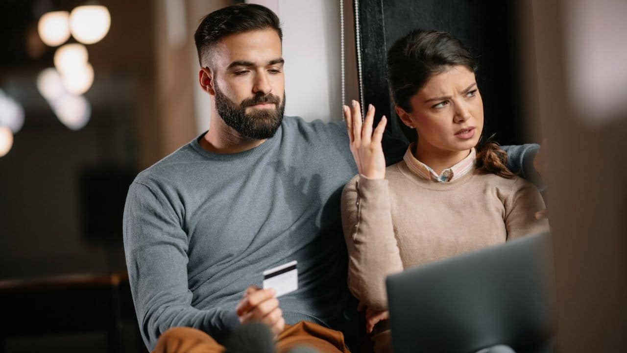A man holds a credit card while a woman sits beside him looking at a laptop.