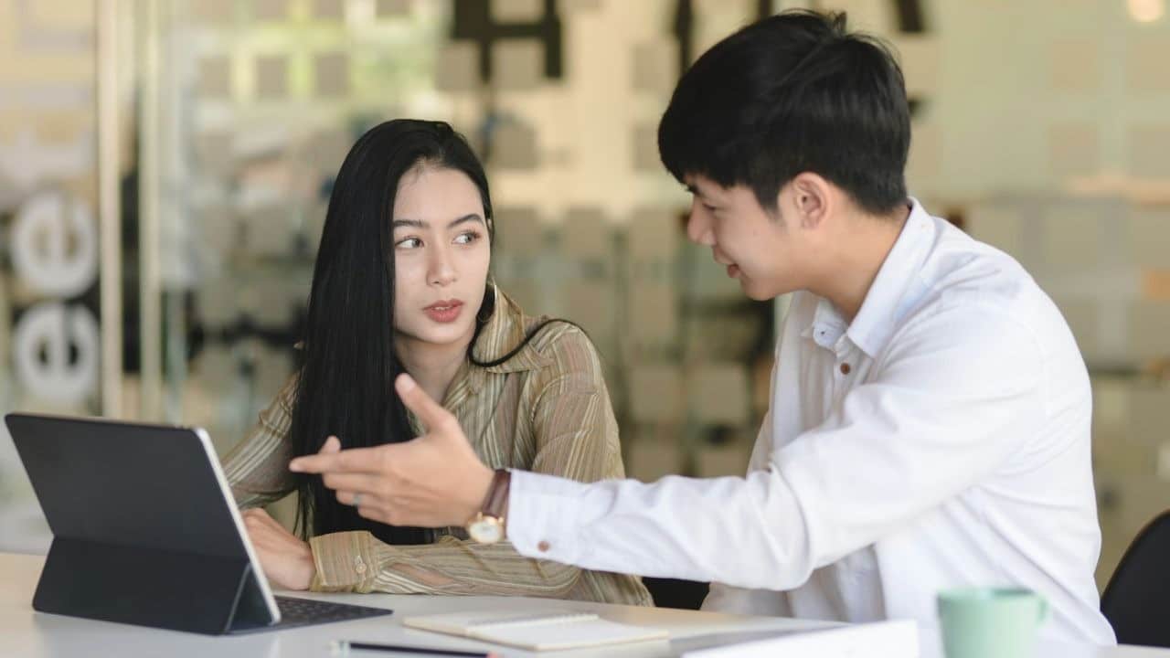 A man gestures toward a tablet screen while speaking to a woman at a desk.