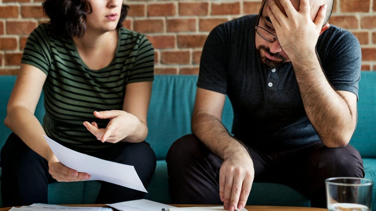 A woman holds papers and gestures while a man sits with his hand on his head.