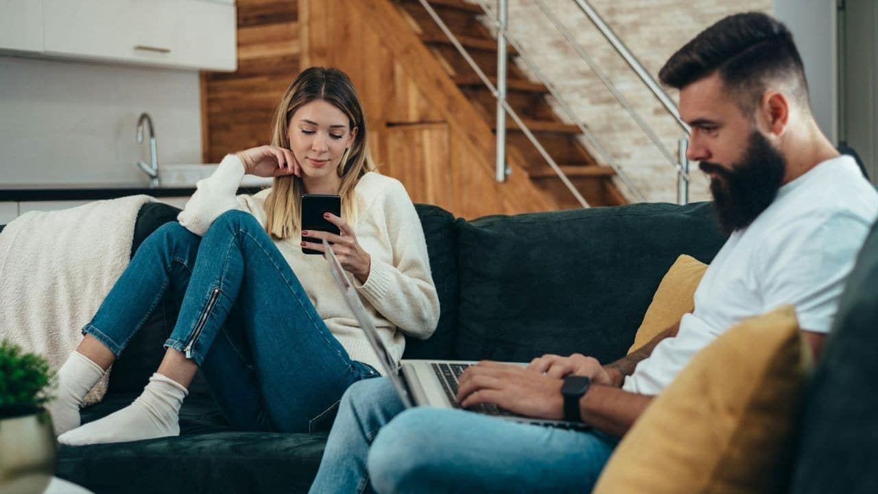 A man uses a laptop while a woman looks at her phone on a couch.