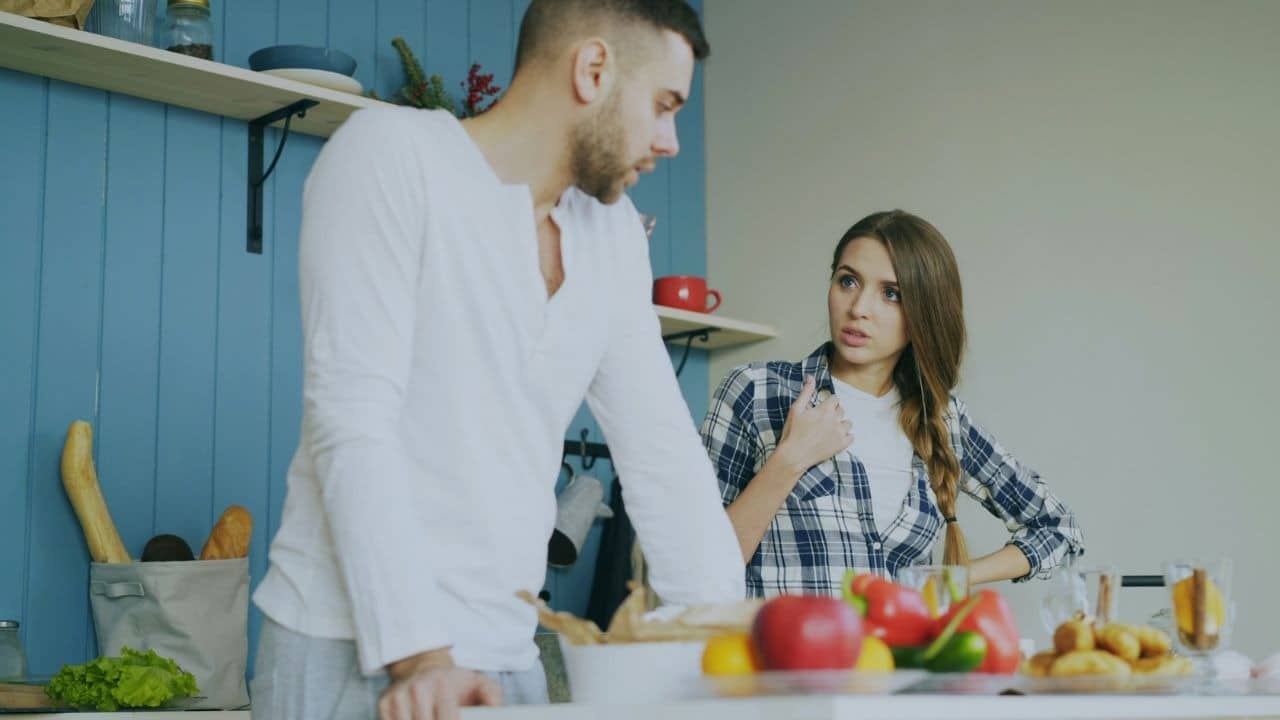 A woman in a plaid shirt speaks to a man while standing in a kitchen.