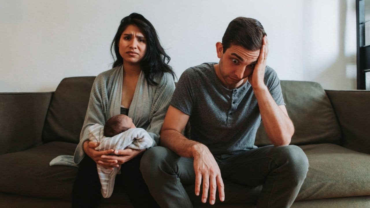 A woman holds a baby while a man beside her sits with his hand on his head.