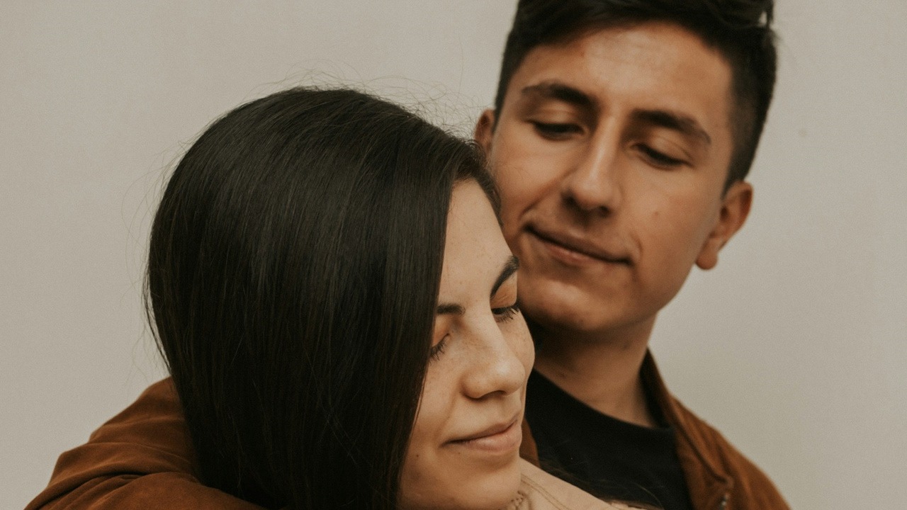 A couple stands close together, with one person embracing the other from behind.
