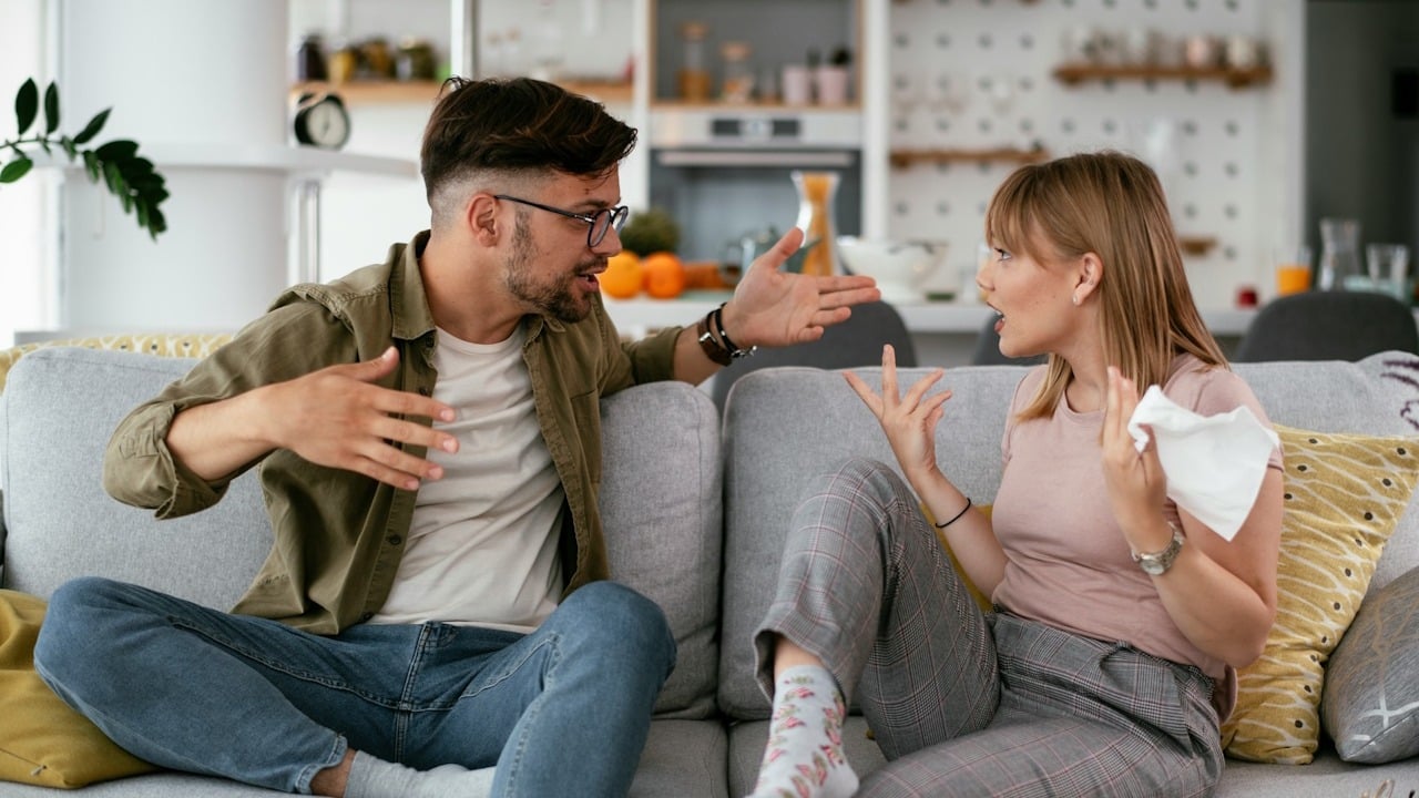 A young couple sitting on a gray sofa, engaged in a heated argument.
