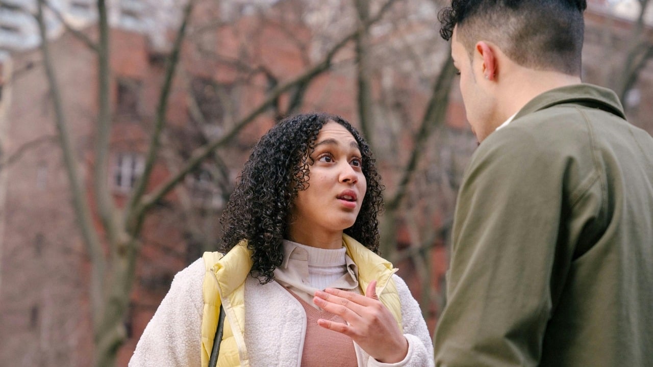 A woman with curly hair looks at a man and gestures while talking outdoors.