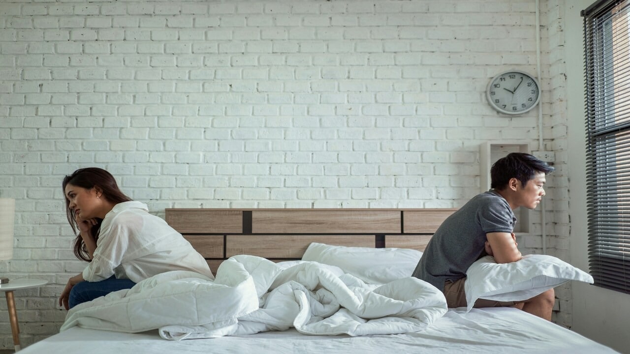 A couple sitting on opposite sides of a bed with their backs against each other.
