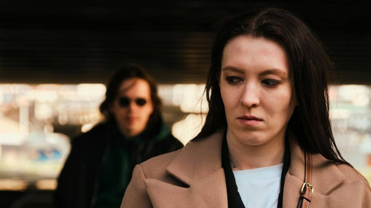Woman in a beige coat looking down, with a blurred person in sunglasses walking behind her.