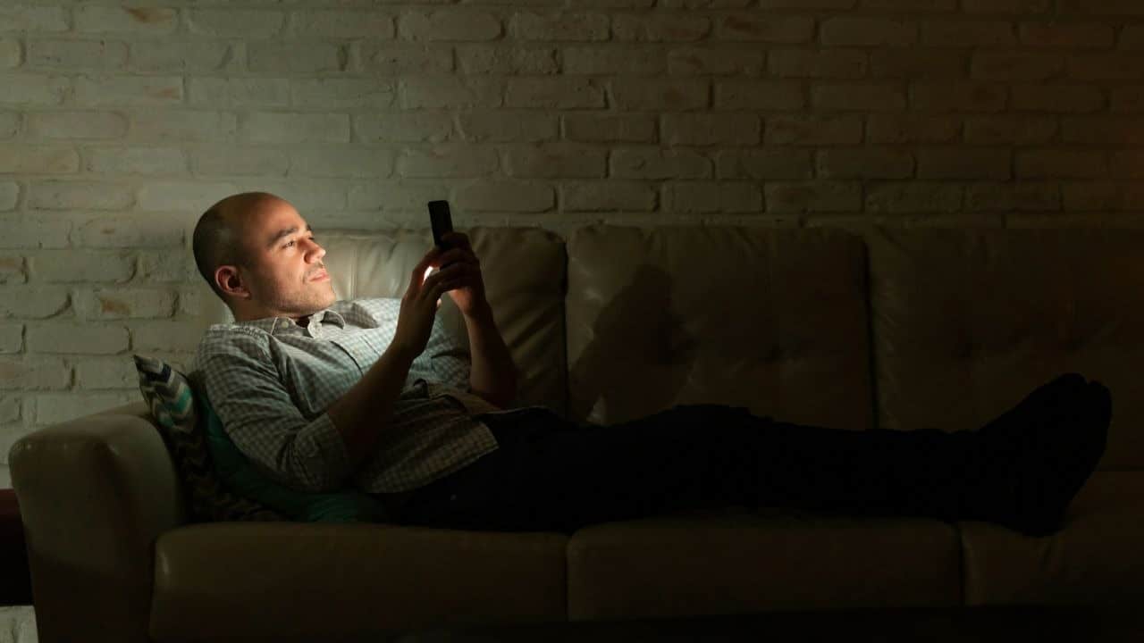 A man lies on a couch in a dark room, illuminated by the bright screen of his smartphone.