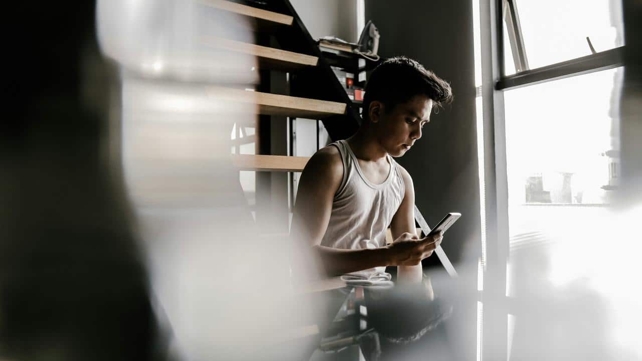 A man sitting by a window under stairs looks down at his phone.