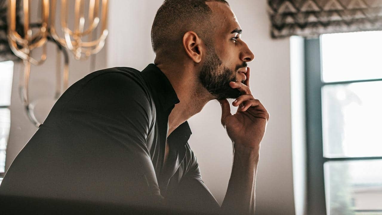 A man sits indoors with his hand on his chin, looking thoughtful.