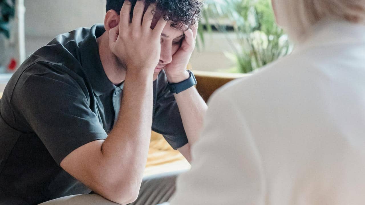 A man sits hunched over, covering his face with his hands in frustration.