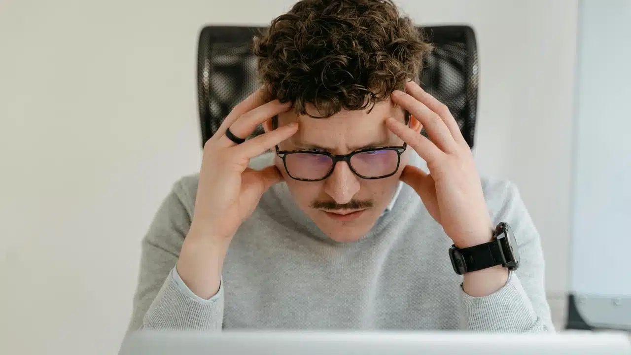 A man with glasses holding his head while looking stressed at a computer.