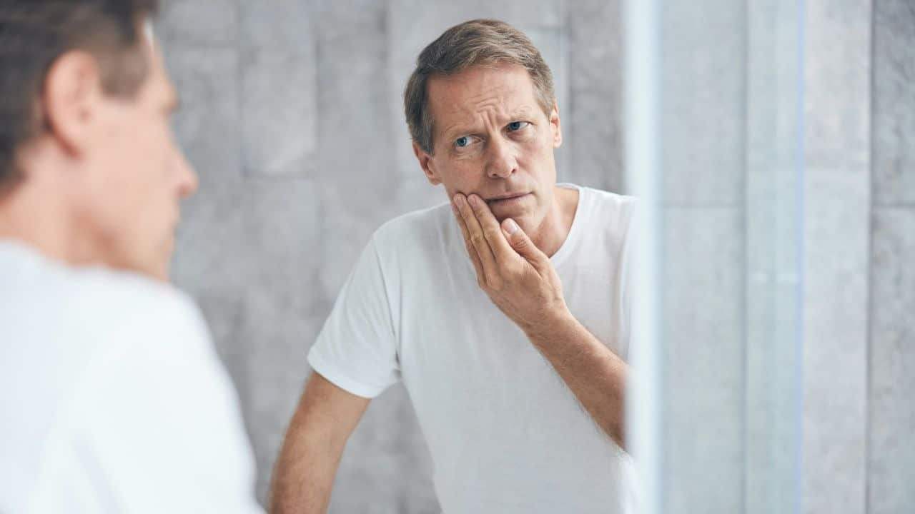 A man in a white t-shirt looks into a mirror while touching his face with concern.