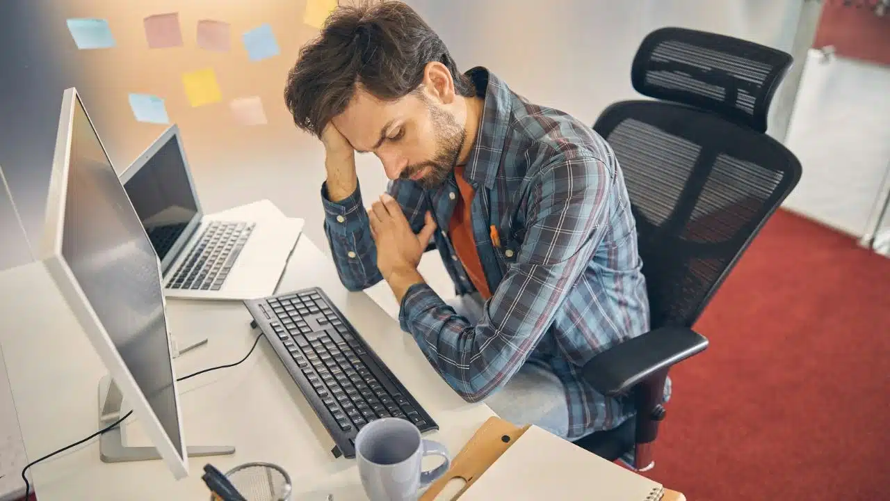 A man in a plaid shirt sits at a desk looking down with his head in his hand.