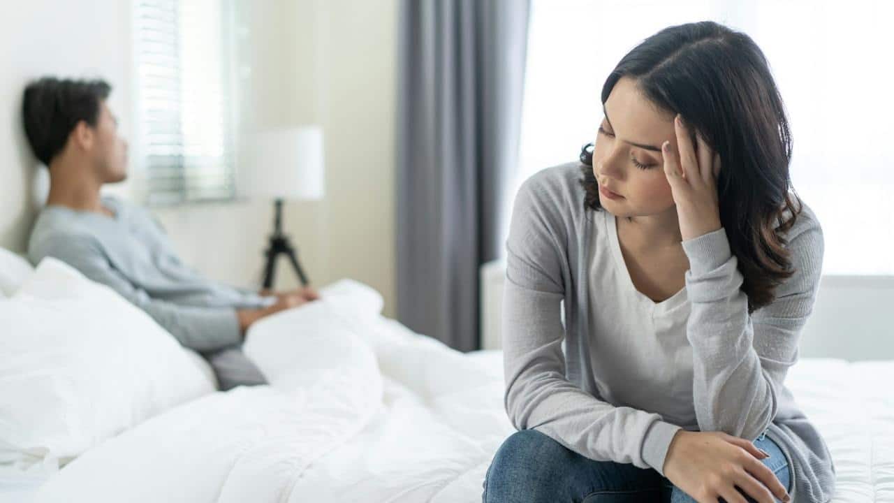 A distressed woman is sitting on the edge of a bed with her head in her hand, while a man sits turned away from her in the background.