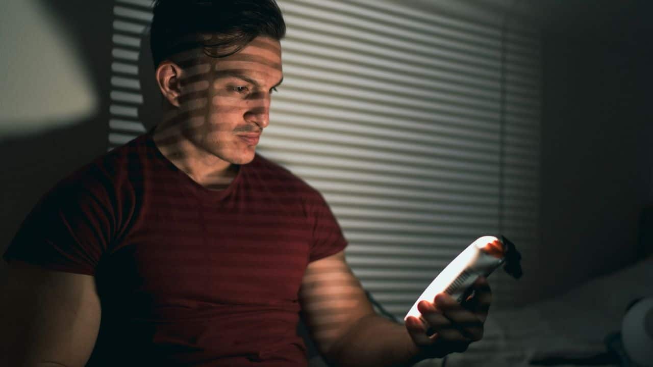 Man in a red shirt looking at a bright phone in a darkened room with blinds.