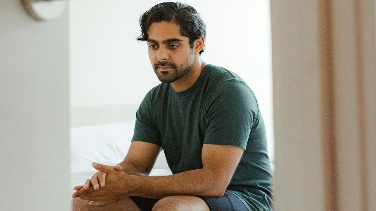 Man with a beard and dark hair sitting indoors, looking down with hands clasped.