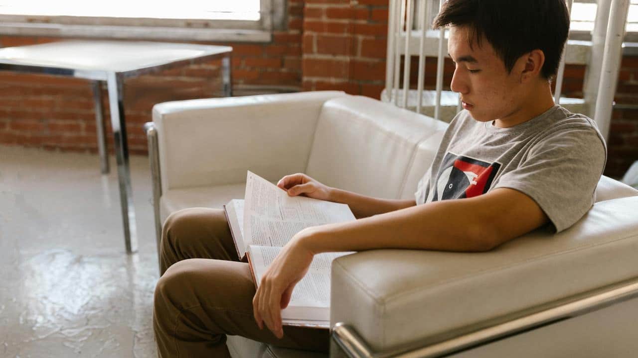 A young man sitting on a couch reading a book.