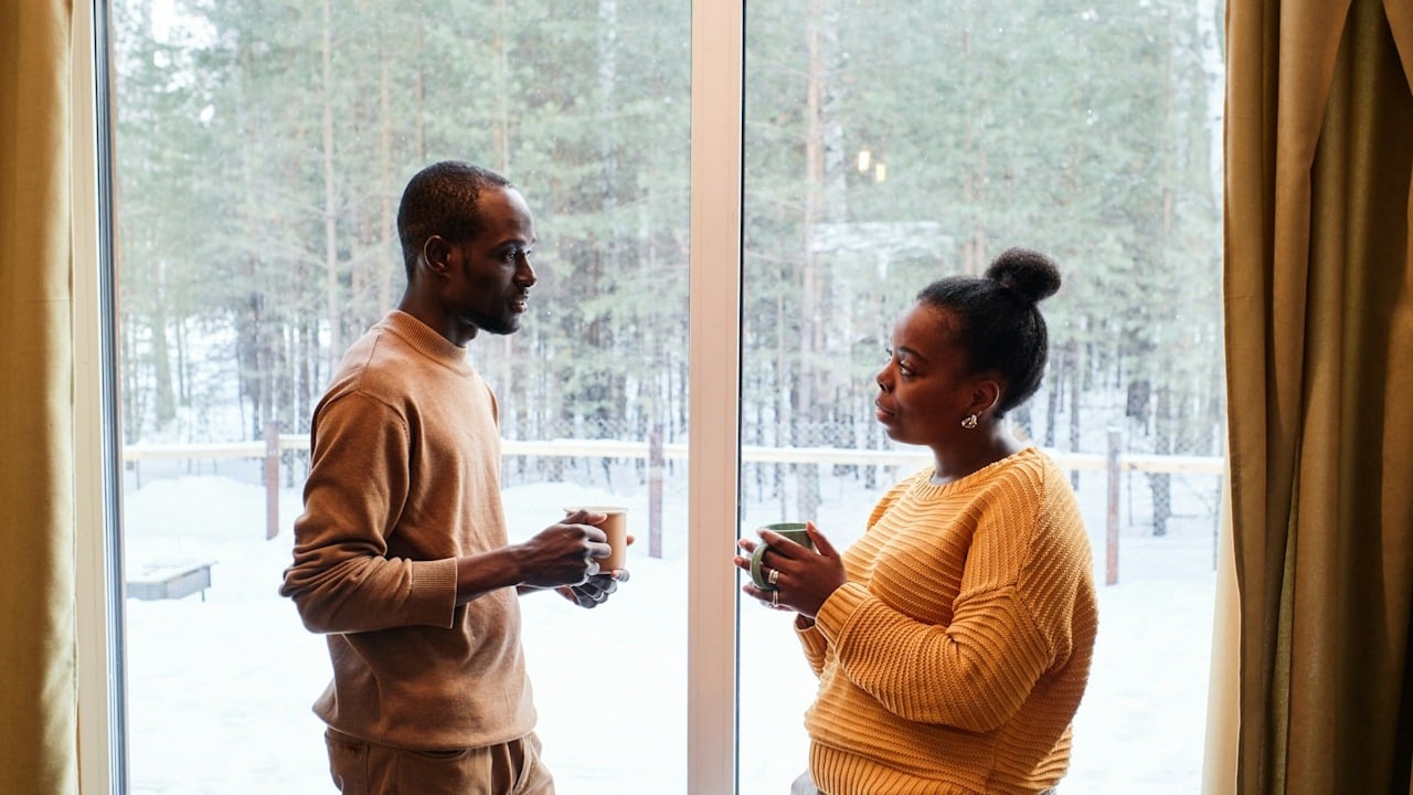 A couple talking and having coffee by a window outside of which there are trees and snow covered ground.