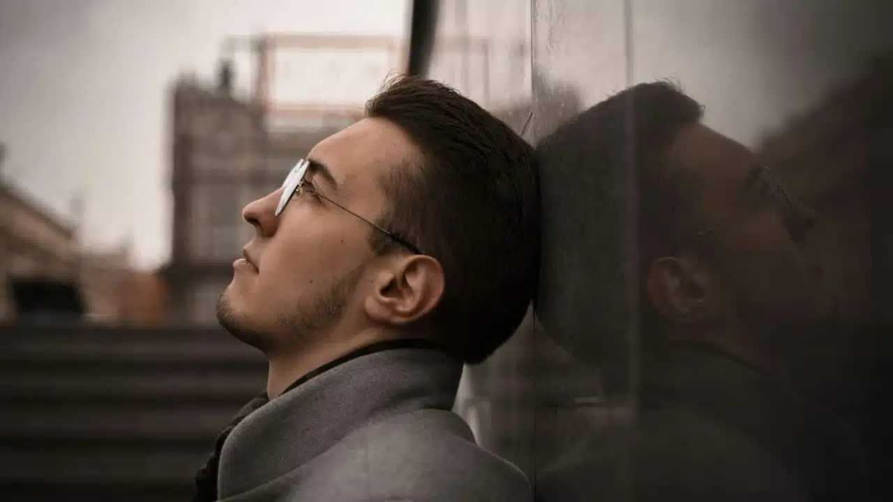 A serious man with glasses and a beard is leaning his head against a stone wall, looking up.