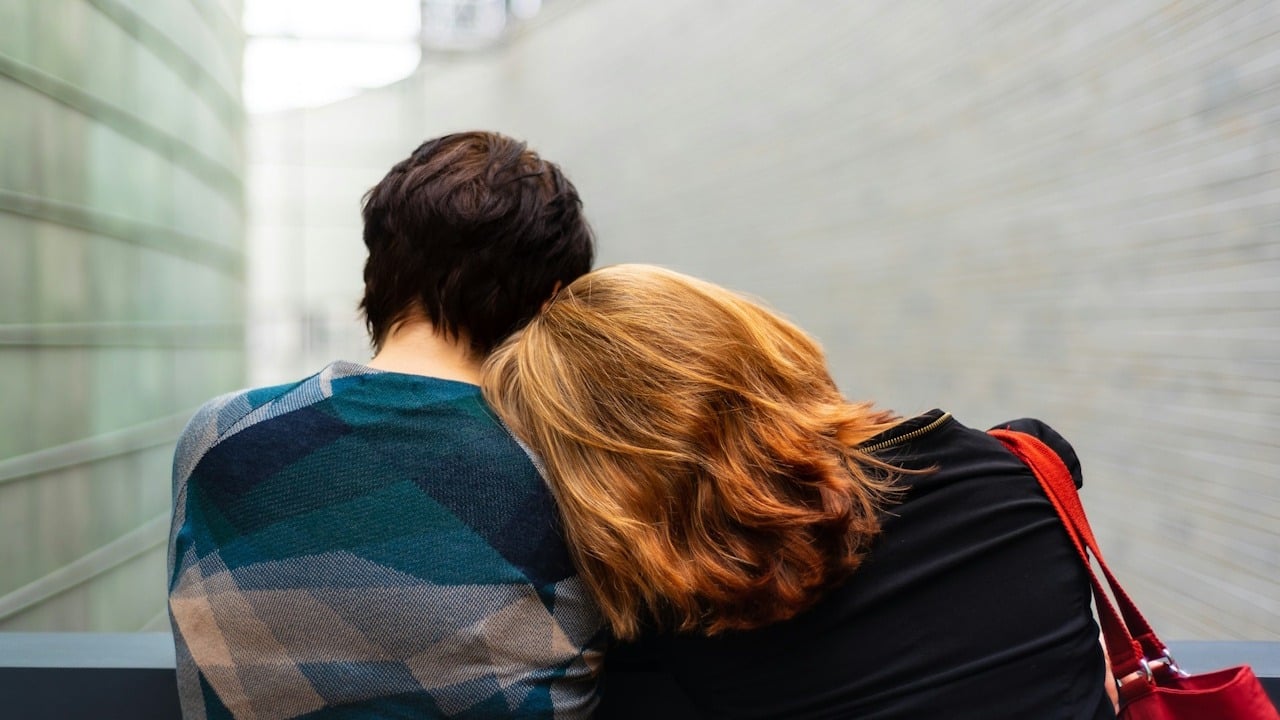Back view of two people, one with their head resting on the other's shoulder.