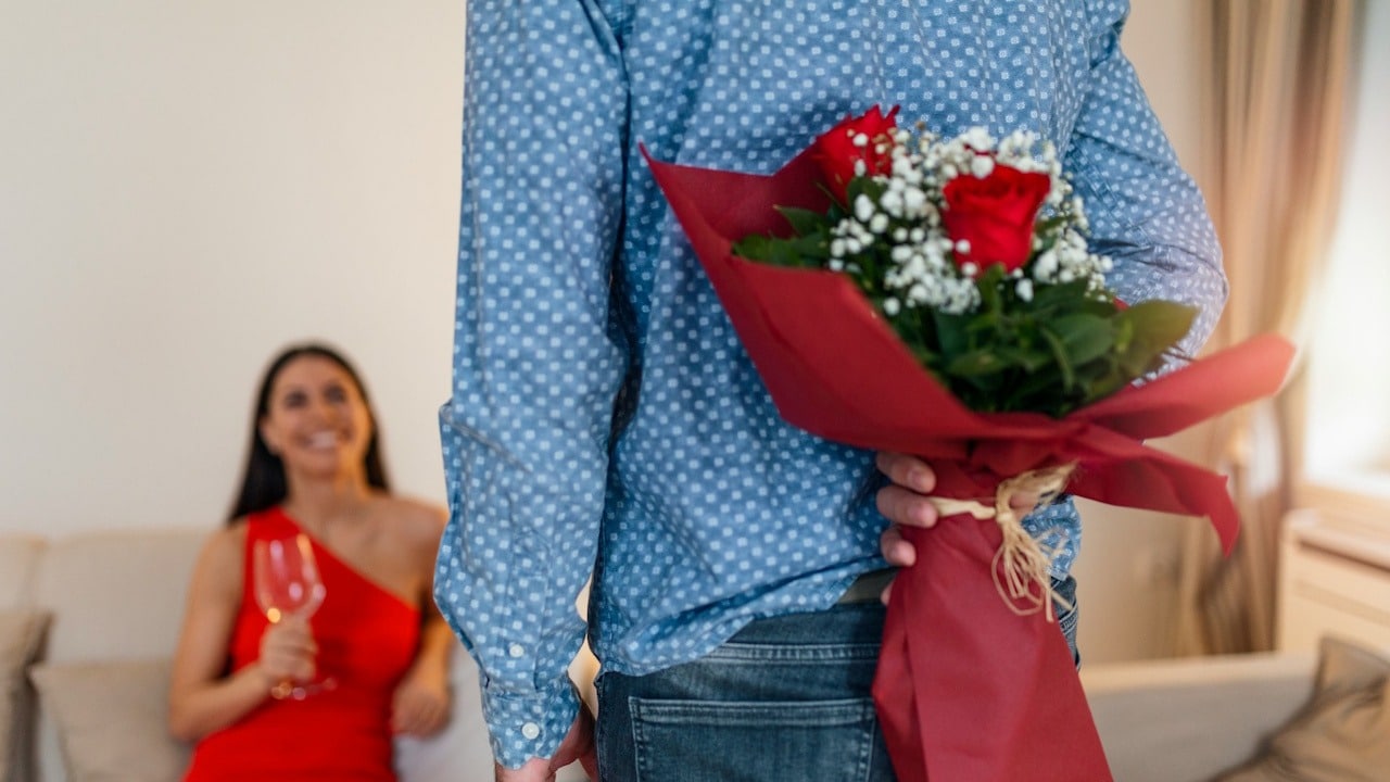 Man holding a bouquet of red roses behind his back for a smiling woman on a couch.