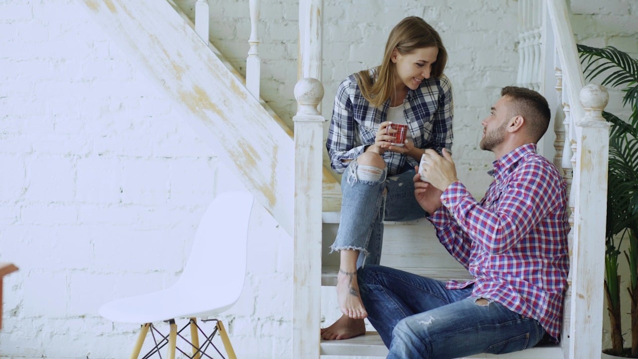 Man and woman sitting on stairs in a bright, modern room, holding mugs and smiling at each other.