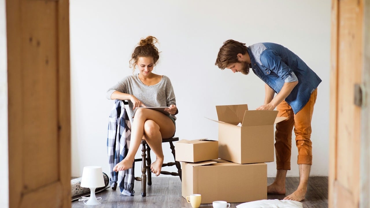 Couple moving into a new home, with the man unpacking cardboard boxes and the woman sitting on a chair looking at a tablet.